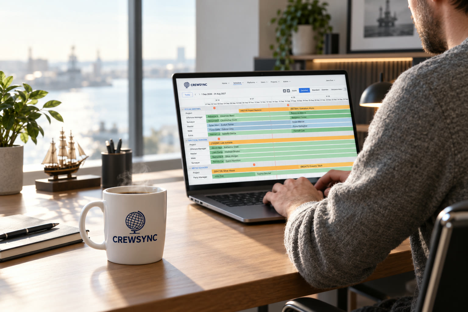 A crewing coordinator working at a desk with a laptop, coffee mug branded with the CrewSync logo beside the keyboard.